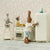 Miniature Maileg kitchen scene with toy rabbit near a refrigerator against a floral wallpaper background