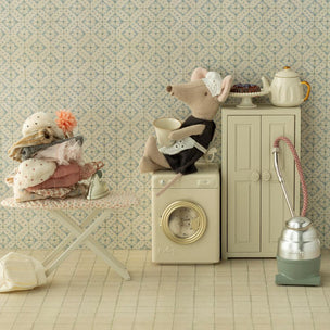 Miniature scene with a Maileg mouse on a washing machine, teapot, and ironing board against a patterned wall.