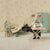 Miniature scene with a stuffed mouse character sitting at a table in a room with floral wallpaper.