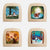 Four square wooden picture frames with rounded corners, arranged in a 2x2 grid on a light gray background. Each frame contains a different painted woodland scene: a girl sitting by ducks, a bear in a glowing forest, a figure among mushrooms, and a couple dancing in a snowy forest.