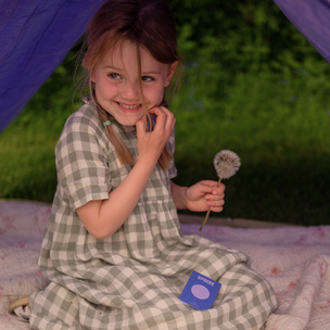 Young girl in a checkered dress sitting inside a tent with a dandelion flower.