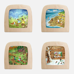 Four square wooden picture blocks with rounded corners arranged in a 2x2 grid on a white background. Each block contains a colorful painted scene: a grassy field with children and animals, a beach with people and boats, a rural landscape with a cabin and figures, and a snowy outdoor scene with birds and a log cabin.
