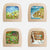 Four square wooden picture blocks with rounded corners arranged in a 2x2 grid on a white background. Each block contains a colorful painted scene: a grassy field with children and animals, a beach with people and boats, a rural landscape with a cabin and figures, and a snowy outdoor scene with birds and a log cabin.