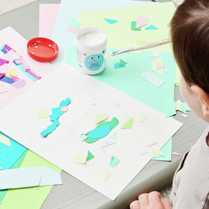 Child engaged in art project with colorful paper shapes and paint on a table.