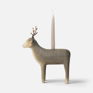 A sculptural Maileg deer-shaped candle holder in a light natural wood finish, featuring a small twig antler detail on its head and a tall white taper candle inserted into its back, set against a plain light gray background.