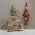 Decorative scene with small tree, gift boxes, and gnome figurines against a plain wall.