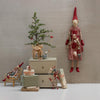 Decorative scene with small tree, gift boxes, and gnome figurines against a plain wall.