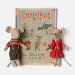 A Maileg children's book titled "Christmas Tales: Bedtime Stories" displayed upright, with two small plush mouse toys positioned in front on both sides. The book cover shows a cozy winter village scene with trees, a house, and red title text.