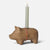 A Maileg wooden pig-shaped candle holder with a lit candle inserted into its back, featuring a red-and-white striped cord and a round wooden charm, photographed against a light gray background.