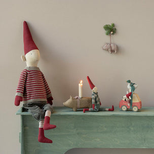 A styled interior scene showing a handmade fabric gnome doll sitting on a painted green shelf against a beige wall. The main doll wears a red and gray striped sweater, gray pants, and red socks, with a tall red hat.