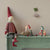 A styled interior scene showing a handmade fabric gnome doll sitting on a painted green shelf against a beige wall. The main doll wears a red and gray striped sweater, gray pants, and red socks, with a tall red hat.