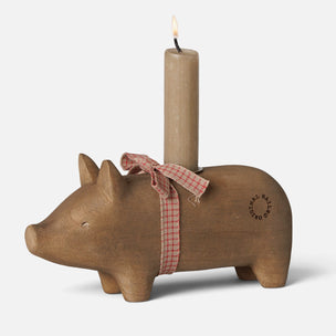 A rustic Maileg wooden pig-shaped candle holder with a lit beige pillar candle inserted into its back. The pig is decorated with a red and white gingham ribbon bow around its neck and features the brand name 'Rustic Hay' printed on its side.