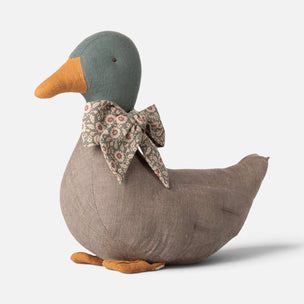 A plush Maileg duck-shaped decorative pillow or toy in textured taupe fabric, featuring a muted green head, orange beak and feet, and a floral-patterned bow tie, photographed on a light gray background.