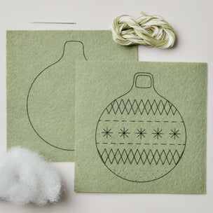 Felt Christmas ornament craft kit with two sage green felt sheets showing outline and embroidered pattern, a bundle of variegated thread, and white stuffing on a light beige background.