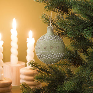 A handmade felt Christmas ornament in sage green hangs on a pine tree branch, decorated with white embroidered geometric patterns and star motifs. Twisted white candles are softly blurred in the background against a warm golden-yellow wall.