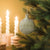 A handmade felt Christmas ornament in sage green hangs on a pine tree branch, decorated with white embroidered geometric patterns and star motifs. Twisted white candles are softly blurred in the background against a warm golden-yellow wall.