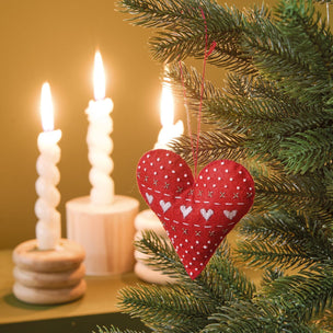 A red fabric heart-shaped Christmas ornament with white embroidered hearts and dots hangs on a green Christmas tree branch, with lit white spiral candles blurred in the background against a warm golden-brown wall.