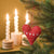 A red fabric heart-shaped Christmas ornament with white embroidered hearts and dots hangs on a green Christmas tree branch, with lit white spiral candles blurred in the background against a warm golden-brown wall.