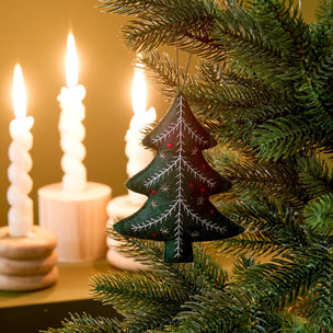 A dark green Christmas tree ornament with white embroidered branches and small red accents hangs on a green artificial Christmas tree branch, with warm, glowing spiral candles blurred in the background.
