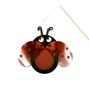 A ladybug-shaped hanging wall decoration made from shiny copper-colored metal sheets, black metal parts, and small green accents, suspended by a string from a thin wooden stick against a plain white background.