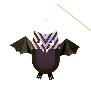 A bat-shaped hanging craft decoration made from black paper wings and body, with a patterned head in lavender, pink, and black, and white eyes, suspended from a wooden stick against a white background.