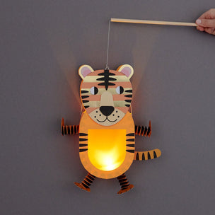 A tiger-shaped hanging paper lantern with a glowing warm light inside, suspended by a string and held by a wooden stick, against a plain gray background. A person's hand is visible at the top right holding the stick.
