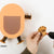 A person's hand is using a hot glue gun to attach a small orange paper paw to a larger orange and brown paper animal craft on a white background.