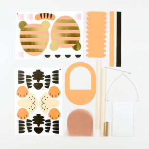 A flat-lay view of a DIY beehive kit containing various components including printed templates of a bee and hive layout, orange and black wood pieces, a wooden frame, a small wooden hive block, and a small wooden stick, all arranged on a clean white background.