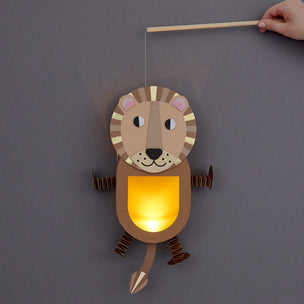 A wooden lion-shaped hanging lantern with a glowing warm yellow light, shown against a dark gray background. A hand holds a wooden stick attached to the top of the lantern.
