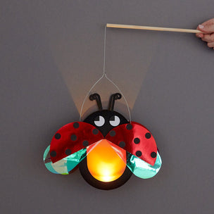 A ladybug-shaped hanging light or lantern made from shiny red and black materials, with a glowing orange center, shown against a plain gray background. A hand holds a wooden stick at the top to manipulate it.