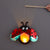A ladybug-shaped hanging light or lantern made from shiny red and black materials, with a glowing orange center, shown against a plain gray background. A hand holds a wooden stick at the top to manipulate it.