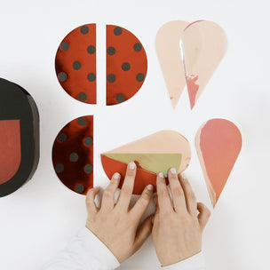 A person's hands are shown assembling a colorful, geometric, circular and teardrop-shaped decorative box or container on a clean white background. The pieces feature metallic finishes in rust red, peach, black polka dots, and olive green.