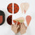 A person's hands are shown assembling a colorful, geometric, circular and teardrop-shaped decorative box or container on a clean white background. The pieces feature metallic finishes in rust red, peach, black polka dots, and olive green.
