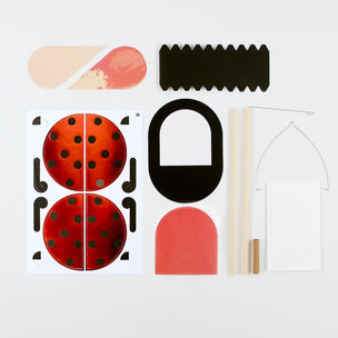 A flat-lay view of a ladybug craft kit containing various colored paper pieces, wooden sticks, and a white sheet on a clean white background.