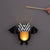 A bat-shaped hanging lantern with black wings and a black-and-white striped face, glowing with warm light from within. A hand holds a wooden stick to swing the lantern against a plain gray background.