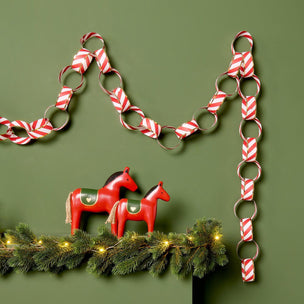 A red and white striped paper chain garland made of paper loops hangs against an olive green wall above a pine garland with warm lights and two red wooden horse figurines.