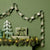 A decorative paper chain garland with metallic silver and green patterned links draped across a dark green wall above a mantel styled with pine garland, a wrapped gift box, and a white tree silhouette.
