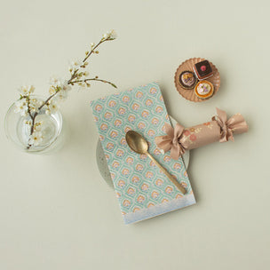 Teal patterned napkin with a gold spoon, crackers, and a small plant on a light green background