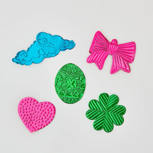 Five colorful decorative shapes made from foil  on a plain light background: a blue cloud, a pink bow, a green patterned egg, a pink heart, and a green four-leaf clover.
