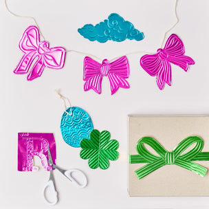 Metallic foil garland set with shiny pink and turquoise bows, a turquoise cloud ornament, a green shamrock, and a green ribbon on a kraft gift box, arranged on a white background with scissors and a pink letter 'R' cutout.