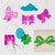 Metallic foil garland set with shiny pink and turquoise bows, a turquoise cloud ornament, a green shamrock, and a green ribbon on a kraft gift box, arranged on a white background with scissors and a pink letter 'R' cutout.