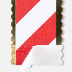 A close-up of a rectangular card or label with a red and white diagonal stripe design and scalloped gold edges, showing a peeled white backing at the bottom on a light gray background.