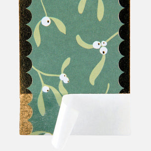 A close-up of a patterned card or notebook cover featuring a green background with mistletoe illustrations, scalloped black border details, and a peeled-back white backing layer.