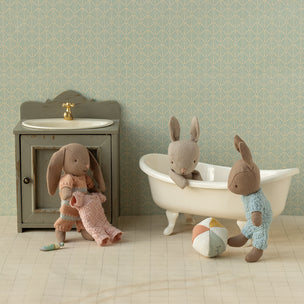 Three plush Maileg rabbit toys in a bathroom setting with a sink, bathtub, and towel.