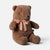 A brown plush Maileg teddy bear sitting down with a dusty pink satin bow tied around its neck, set against a plain light gray background.