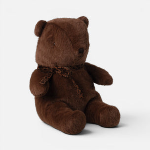 A brown plush Maileg teddy bear sitting upright, wearing a patterned brown and black ribbon bow around its neck, set against a plain light gray background.