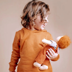 A child wearing a rust-orange long-sleeve top holds a doll with orange hair and an orange outfit against a plain beige background.