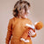 A child wearing a rust-orange long-sleeve top holds a doll with orange hair and an orange outfit against a plain beige background.