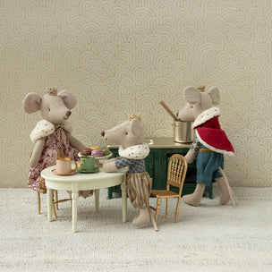 Four Maileg toy mice with various outfits sitting around a small table against a patterned wall.