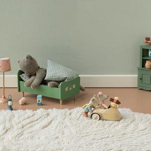 A styled children's bedroom scene with a green wooden bed frame holding a gray stuffed bear and pillows, a pink lamp, a small green cabinet, and a wooden mouse toy driving a car on a white shag rug against a pale green wall.
